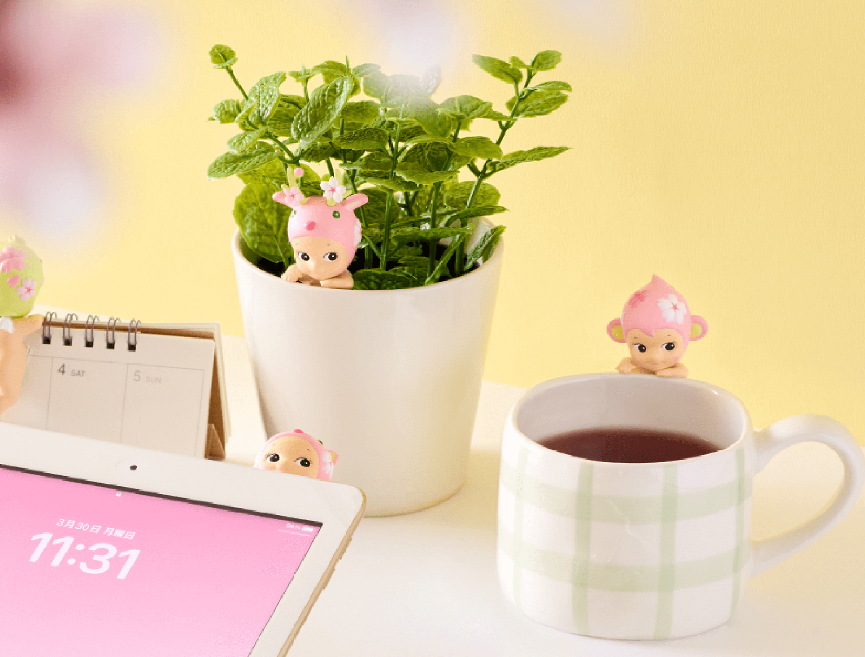 Table with tablet, mug of tea, and small plant with decorative sonny angel figures on a yellow background